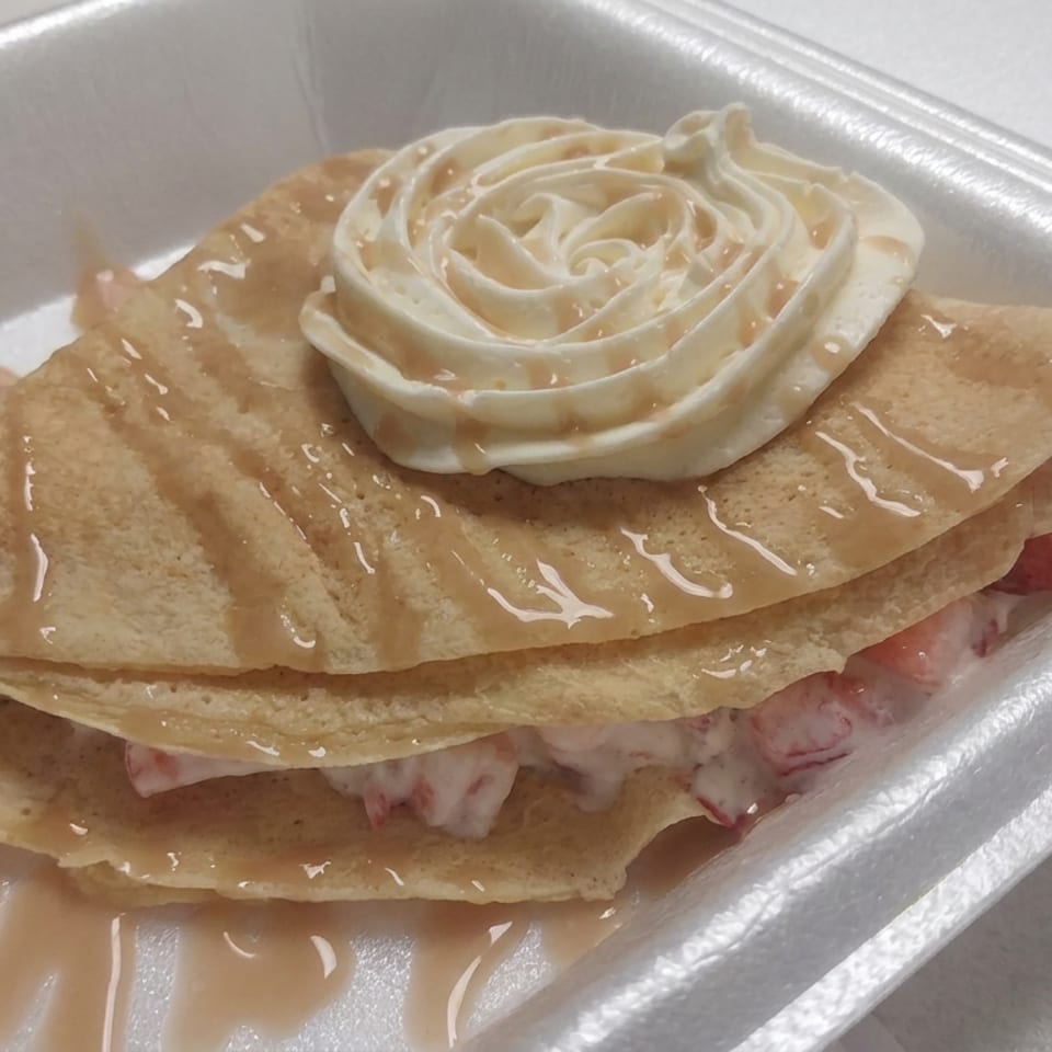 Folded golden crepe filled with creamy fruit, drizzled with caramel sauce and topped with whipped cream in a foam container.
