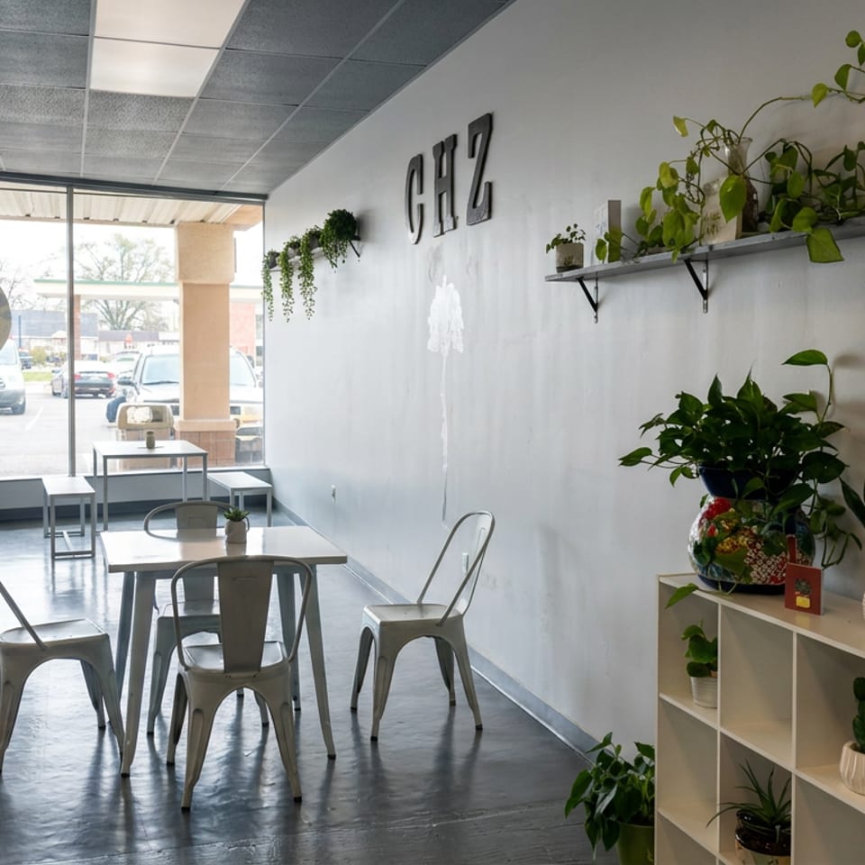 Bright restaurant dining area with metal tables, chairs, abundant green plants, large windows, and wall lettering.