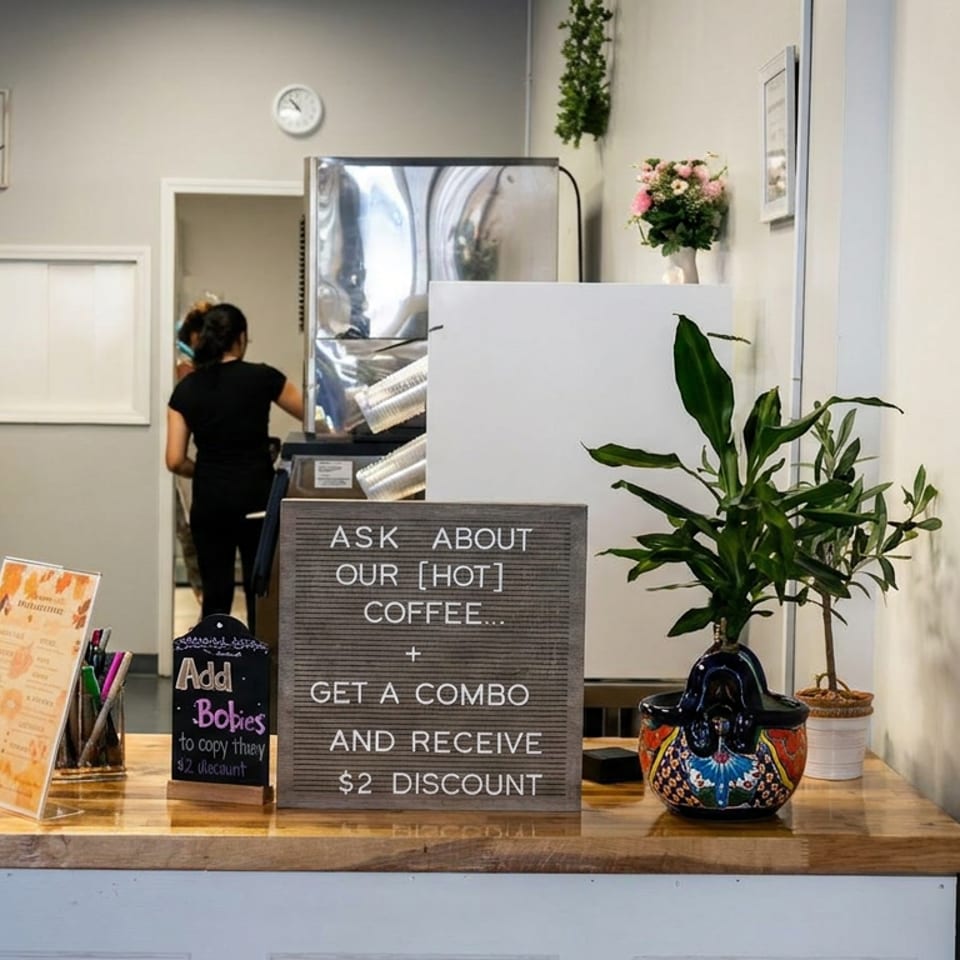 Café counter displays promotional signs, potted plants, register, beverage equipment, and a worker serving customers.