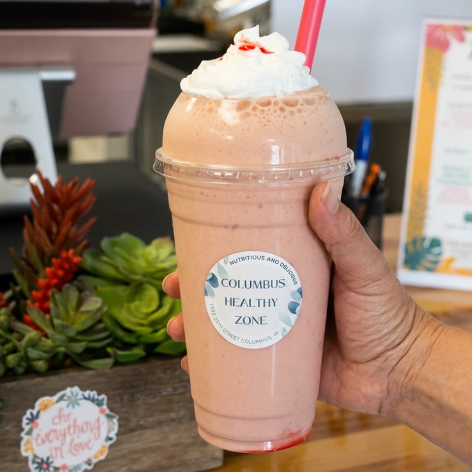 Hand holding a whipped-cream-topped pink smoothie with a straw and Columbus Healthy Zone label inside a restaurant.