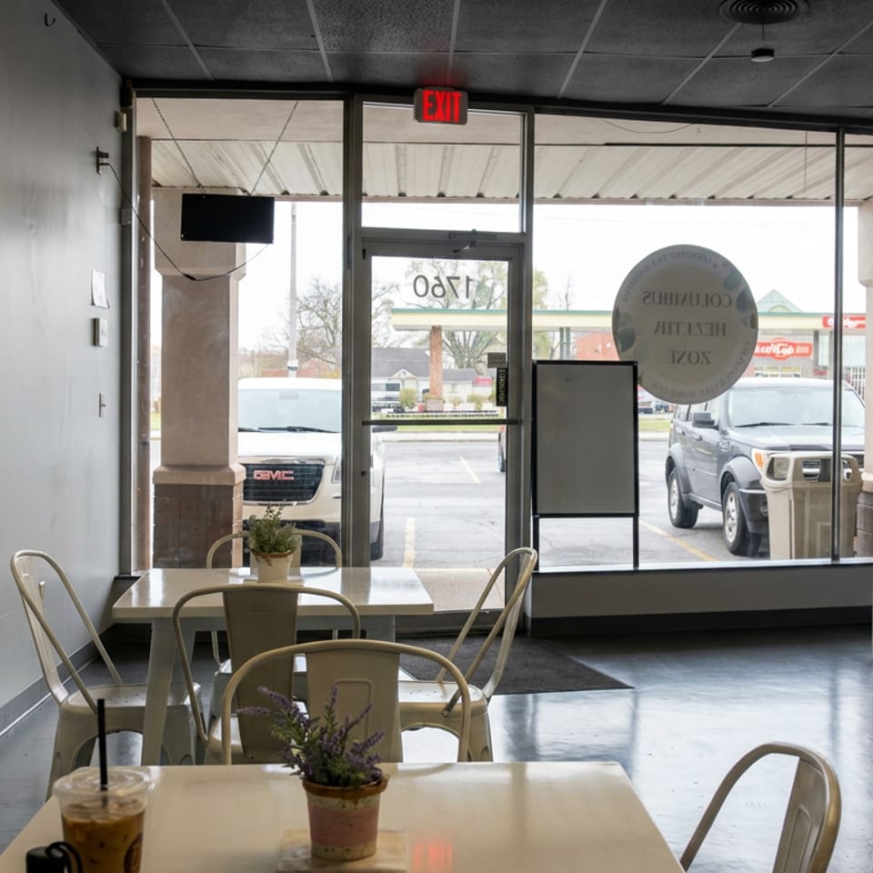 Bright minimalist café interior with metal chairs, tables, potted plants, and floor-to-ceiling windows overlooking a parking lot.