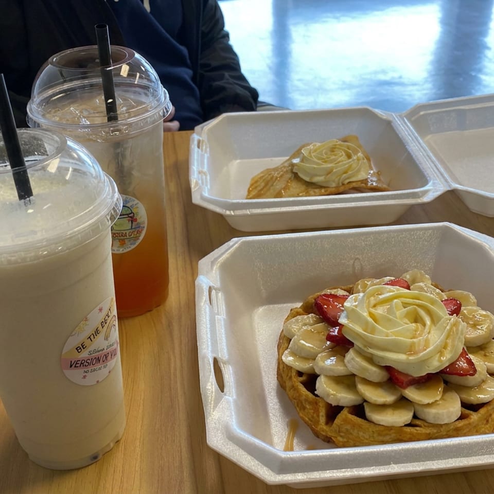 Waffles topped with bananas, strawberries, and whipped cream sit beside two iced drinks in takeout containers.