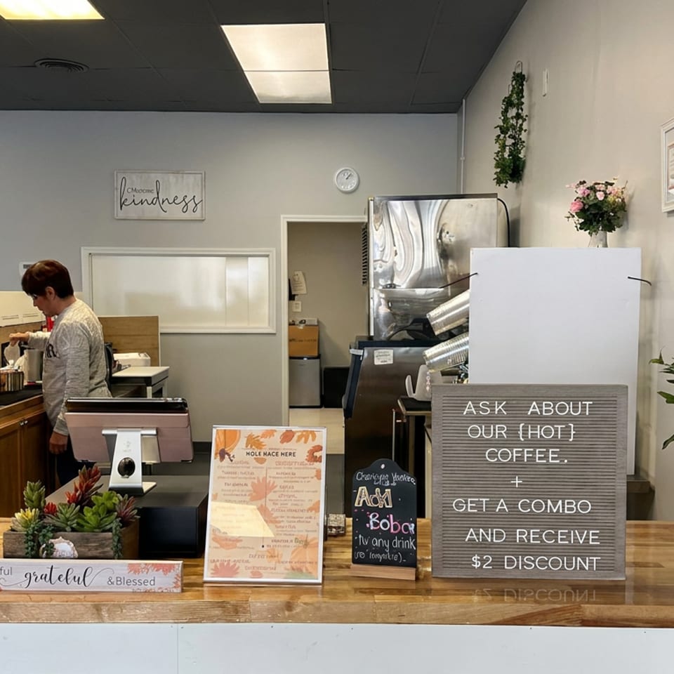 Cozy café counter with pastry display, espresso machine, menu signs, hot coffee promotion, plants, and a staff member.